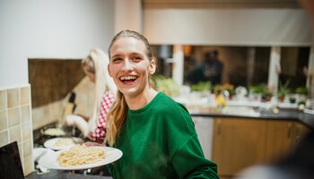 Lachendes Mädchen steht in der Küche und hat zwei Teller mit Nudeln in den Händen | © DGLimages - Getty Images/iStockphoto