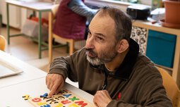 Ein älterer Mann versucht Buchstaben aus Holz in eine dafür vorgesehene Einkerbung zu legen | © Caritas Werkstatt für Menschen mit Behinderung München