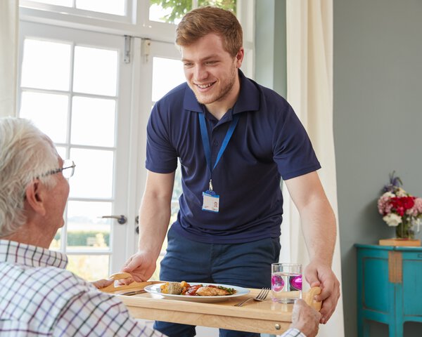 Betreuer bringt Senior Essen auf einem Tablett | © Monkey Business Images - Shutterstock
