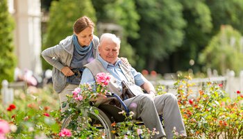 Pflegekraft und älterer Mann im Rollstuhl gehen im Park spazieren | © FredFroese - iStock