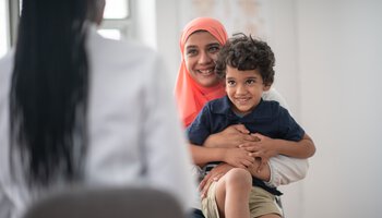 Eine muslimische Mutter ist mit ihrem Sohn beim Arzt, um sich untersuchen zu lassen. Der Junge sitzt auf dem Schoß seiner Mutter und sie umarmt ihn ganz fest. | © FatCamera